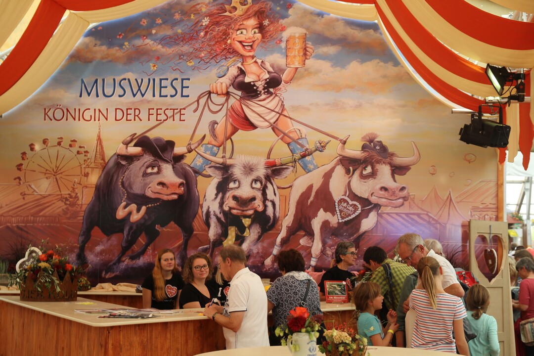 Das Muswiesenmotto 2018: Königin der Feste. Das Wandbild am Gemeindestand im Gewerbezelt hat der Künstler Koval humorvoll entworfen und umgesetzt. Es zeigt die Muswiese als attraktive Volksfestkönigin, die ihre tierischen Rivalen Fränkisches Volksfest (Crailsheim), Cannstatter Wasen (Stuttgart) und Oktoberfest (München) souverän im Zaum hält.